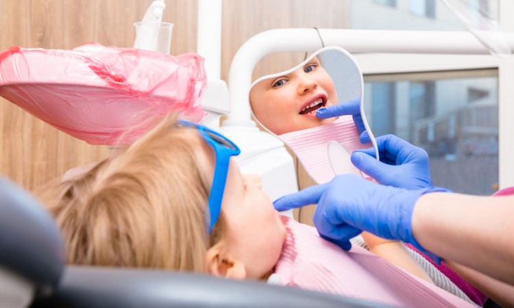 Dentist showing to little girl cured tooth in pediatric dental clinic. Child is sitting in a dental chair inspecting her teeth looking thru tooth-shaped mirror