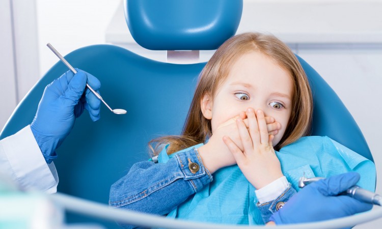 Little cute girl is closing her mouth with hands, preventing examination of teeth, because of fear of doctor. Child is sitting in chair in dental clinic, office. Visiting dentist with children.